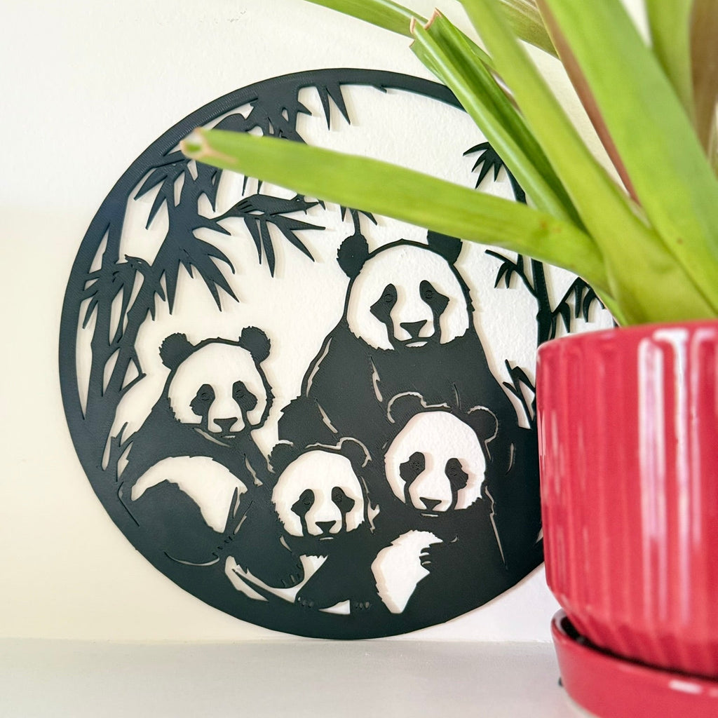 A circular black 3d printed wall art piece featuring a panda family silhouette, styled next to a green potted plant on a white surface against a white wall.
