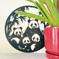 A circular black 3d printed wall art piece featuring a panda family silhouette, styled next to a green potted plant on a white surface against a white wall.