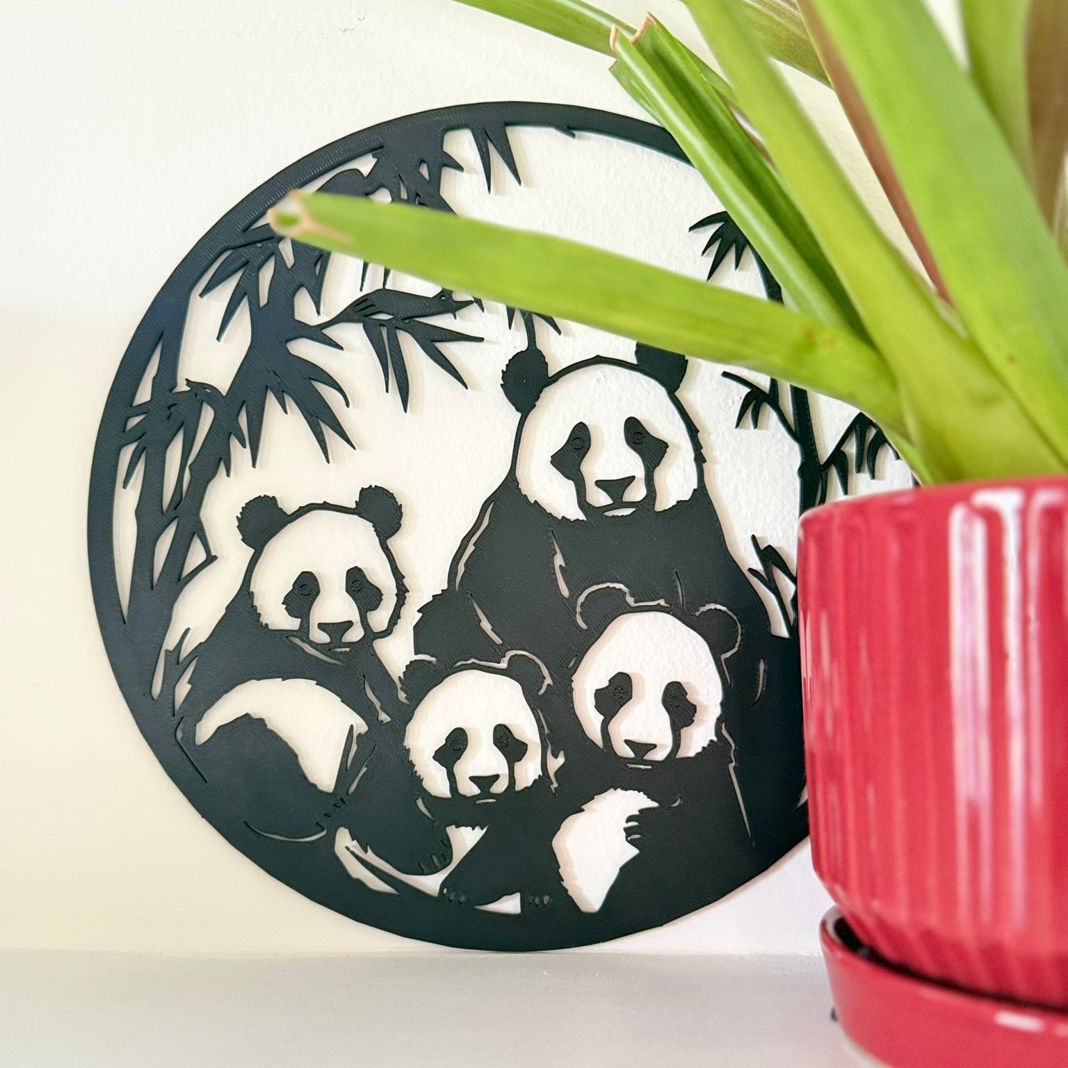 A circular black 3d printed wall art piece featuring a panda family silhouette, styled next to a green potted plant on a white surface against a white wall.