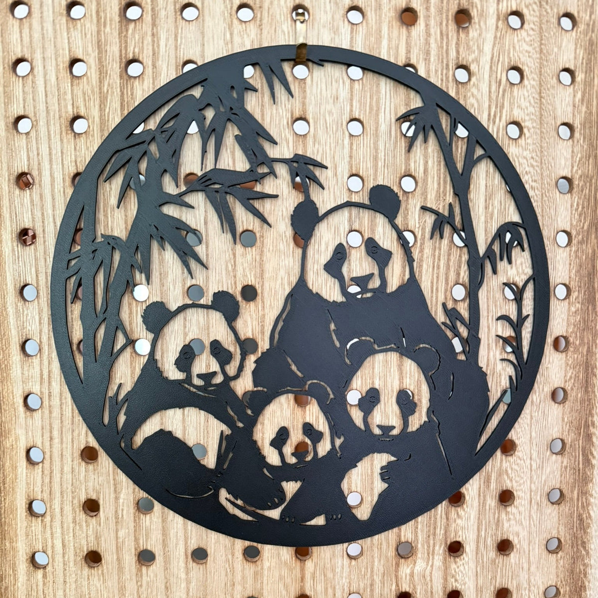 A circular black 3d printed wall art piece featuring a family of pandas and bamboo leaves, mounted on a light wood pegboard background.