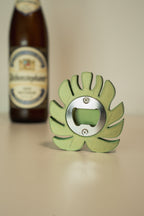 Green bottle opener with leaf design in front of a beer bottle on a beige surface