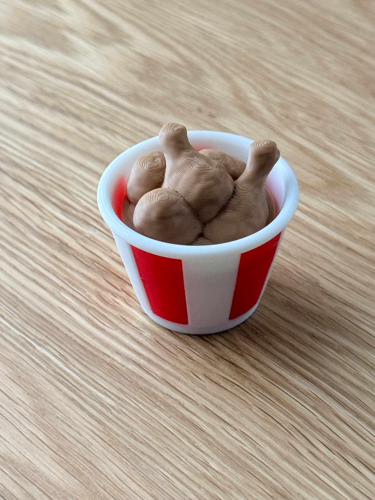 A miniature white and red striped bucket filled with brown toy chicken legs, placed on a light wood grain surface.