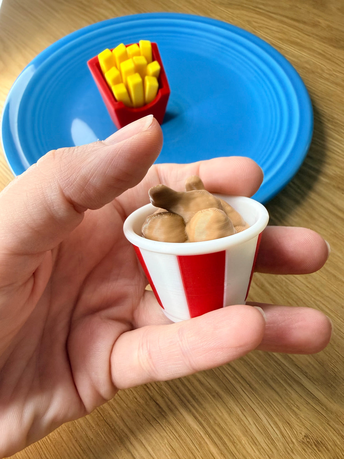 A hand holding a miniature red and white striped cup filled with brown toy food, with a blue plate and yellow toy fries in the background on a wooden table.