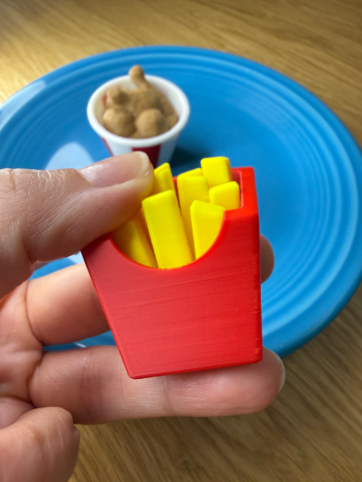 A miniature red and yellow plastic toy french fries container held in a person's hand, with a blue plate and small toy cup in the background.