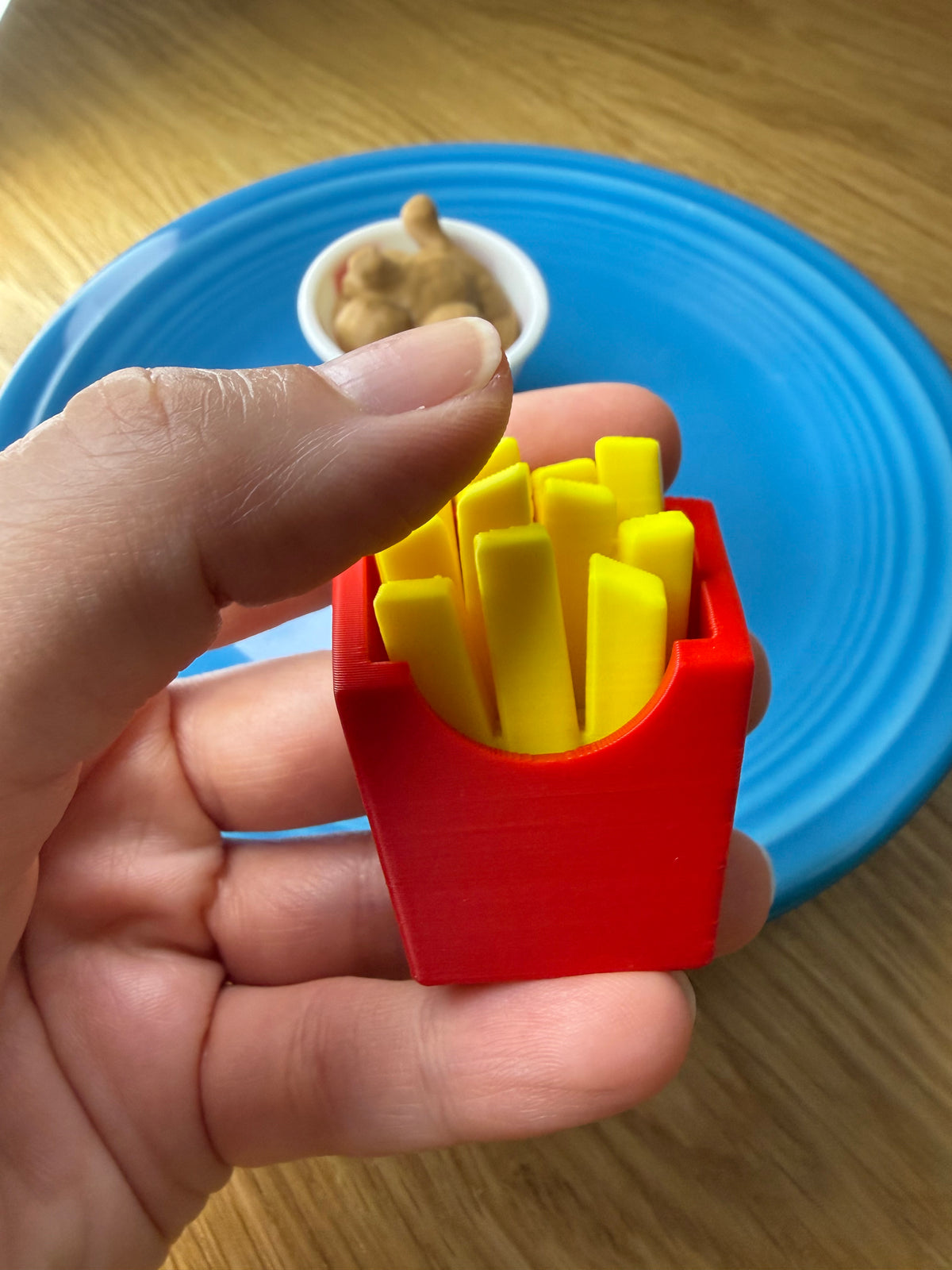 A hand holding a small red container filled with yellow french-fyze-like pieces, with a blue plate and a small bowl in the background on a wooden surface.