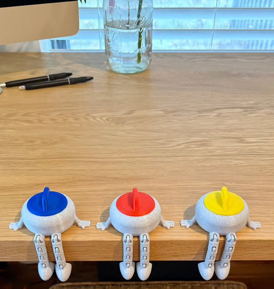 Three colorful curling stone buddies on a wooden desk with pens and a glass of water in the background.