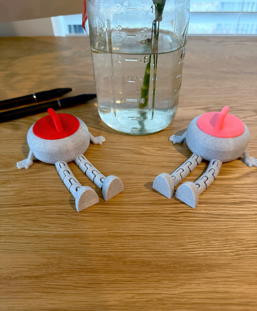 Two small flexi curling stone buddies with red and pink lids on a wooden surface next to a glass of water.